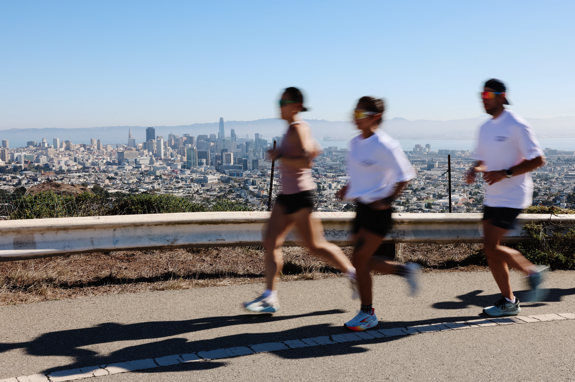 Three runners in Running Wylder gear jogging along a hilltop road with panoramic views of the San Francisco skyline
