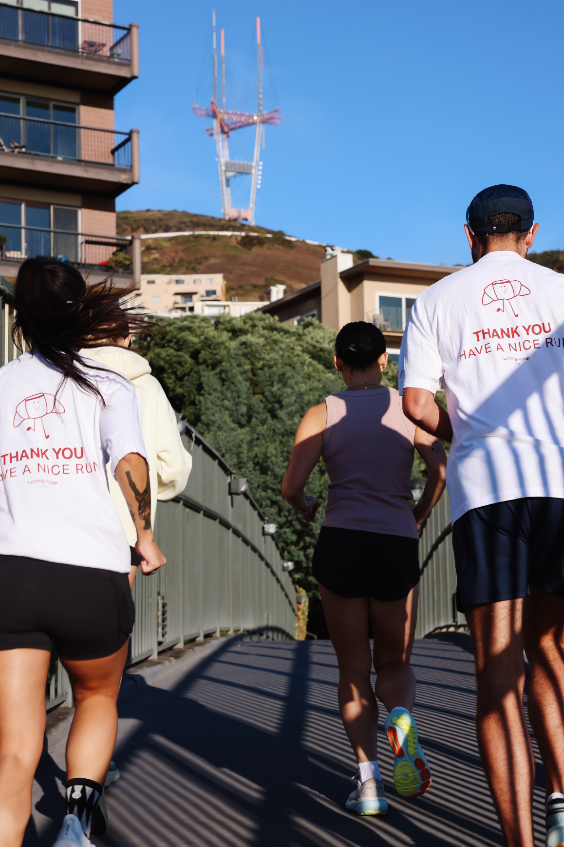 Three runners in Running Wylder apparel crossing a sunlit San Francisco bridge toward Sutro Tower