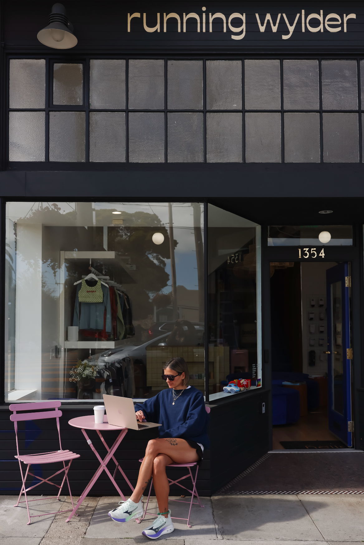 Woman working on a laptop at a pink café table outside the Running Wylder store at 1354 Castro Street, with apparel displayed in the window behind her