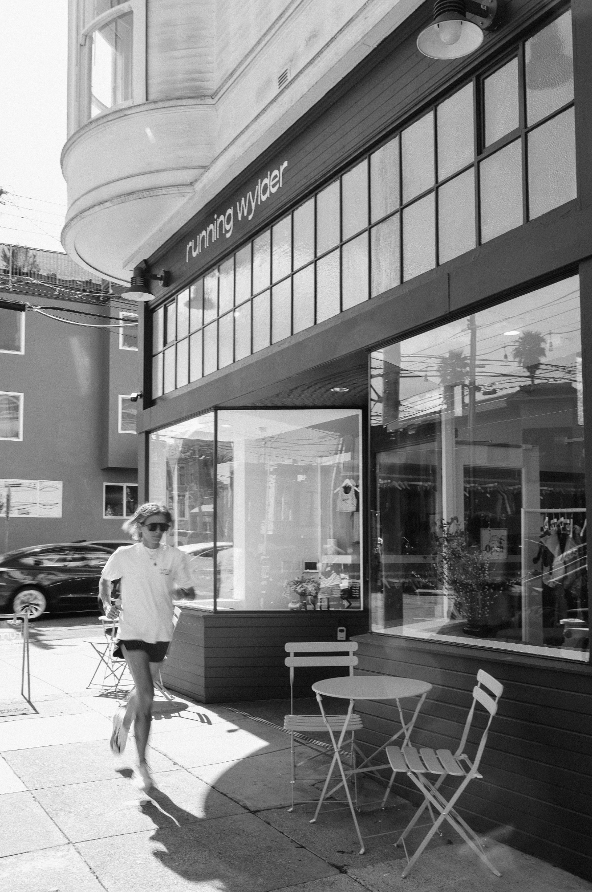 Black and white photo of a runner passing by the Running Wylder storefront with outdoor seating and display windows