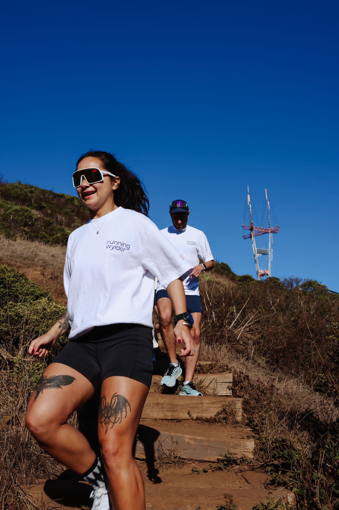 Two runners in Running Wylder gear descending a dirt trail on Twin Peaks with Sutro Tower in the background